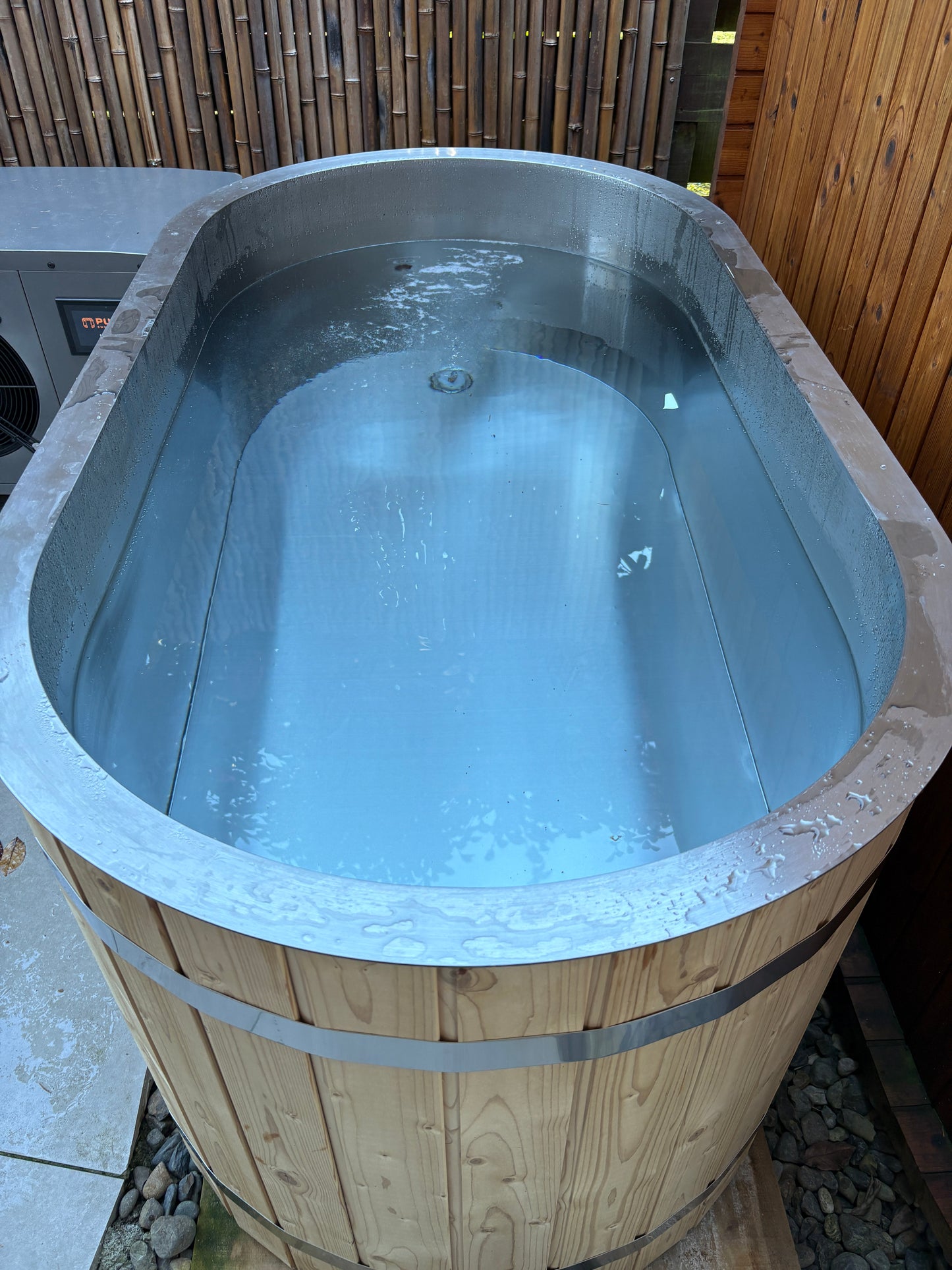 Wooden ice bath with metal interior, surrounded by a wooden deck and stone wall.