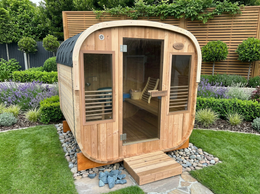 Wooden outdoor sauna in a garden setting with greenery and a wooden fence.