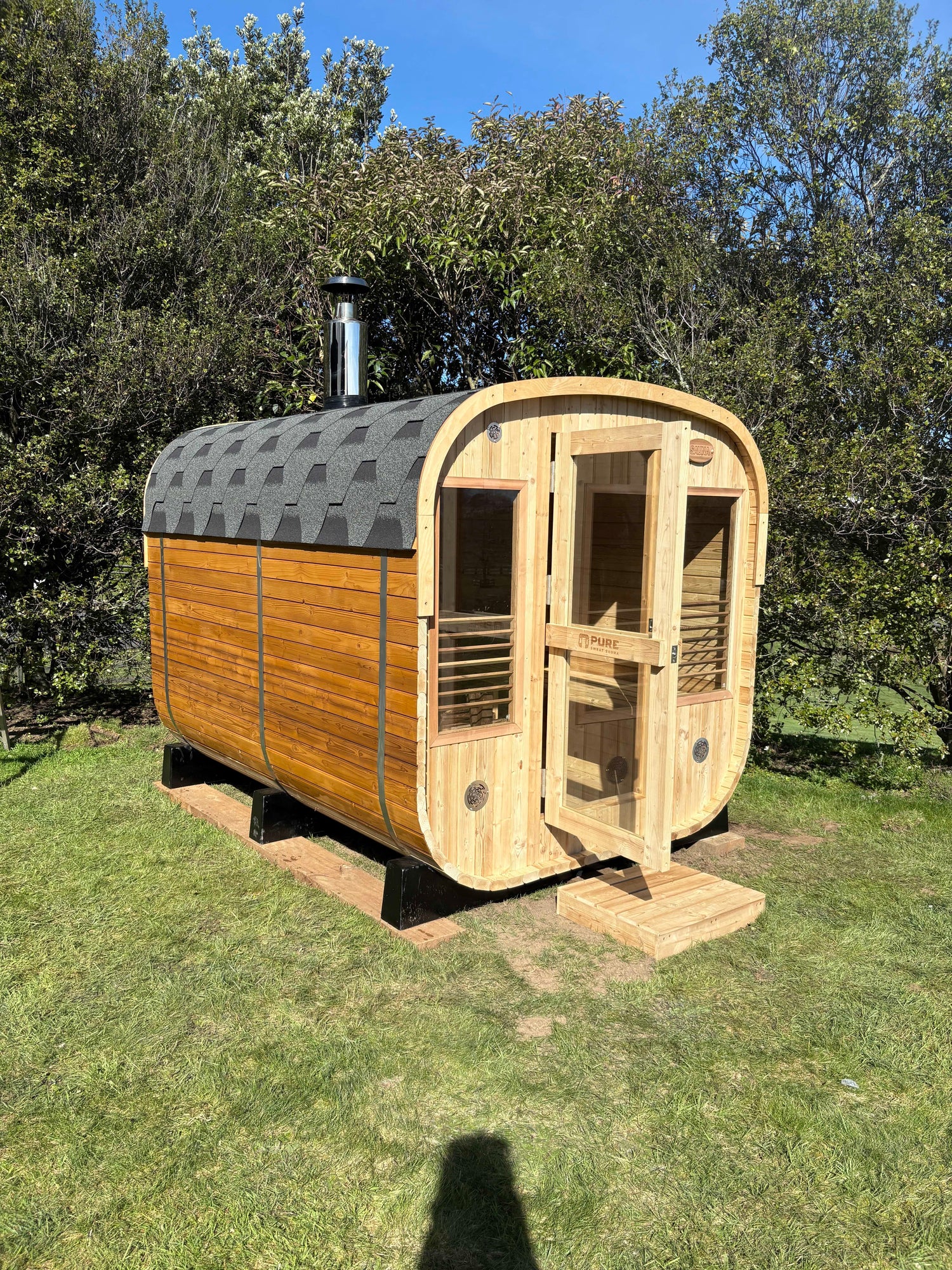 Wooden sauna with a gray roof in a grassy area with trees in the background